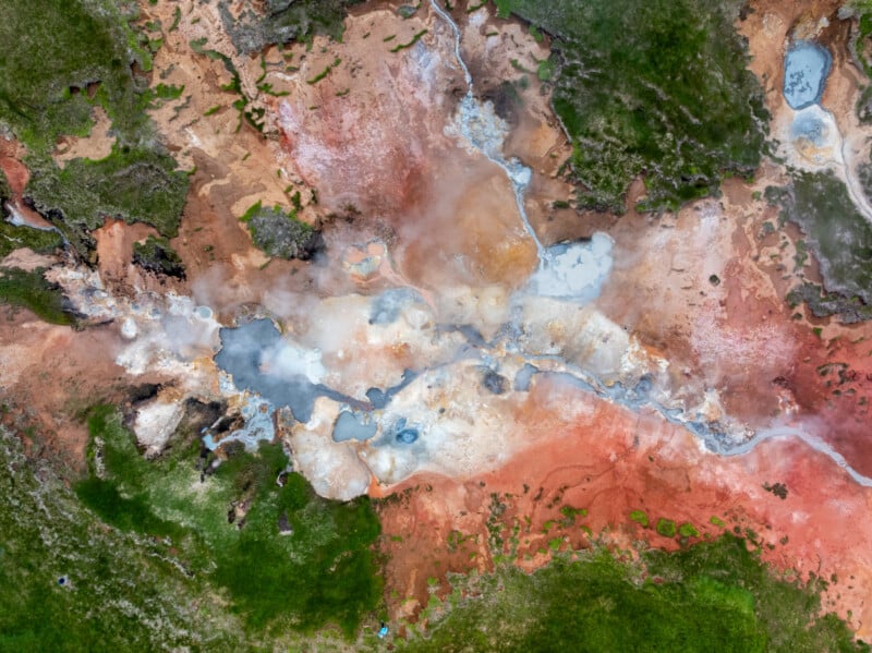 Aerial view of a geothermal area with vibrant mineral deposits in red, orange, and white, steaming hot springs, and patches of green vegetation surrounding the colorful landscape.