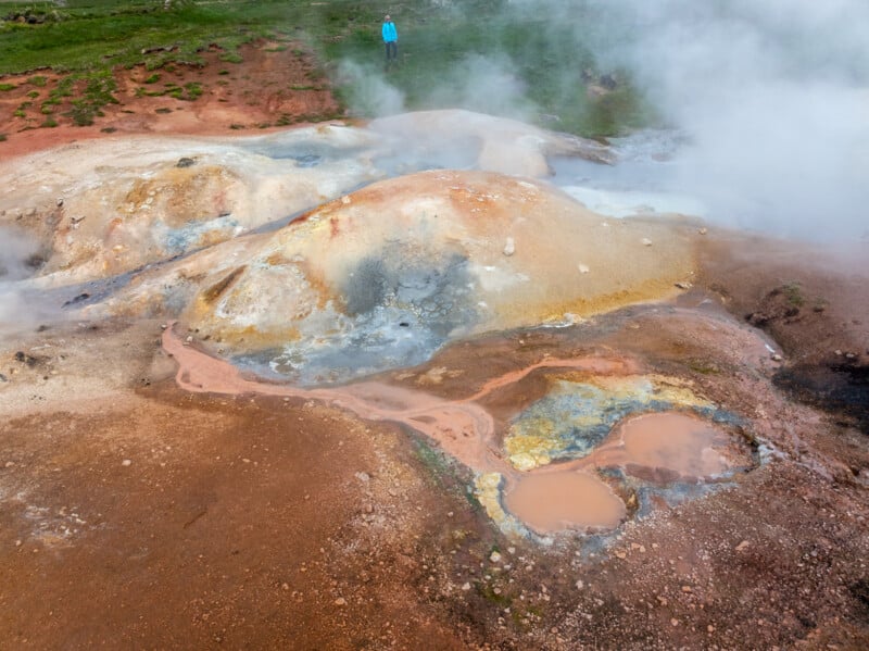 A person stands near colorful geothermal hot springs with steaming vents, mineral-stained ground, and warm pools surrounded by green grass.