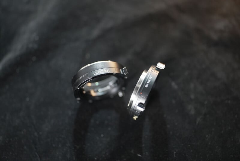Two metallic camera lens adapters are placed on a dark, soft surface. One adapter stands upright while the other leans at an angle. The adapters have visible connection points and some text engraved on their surfaces.