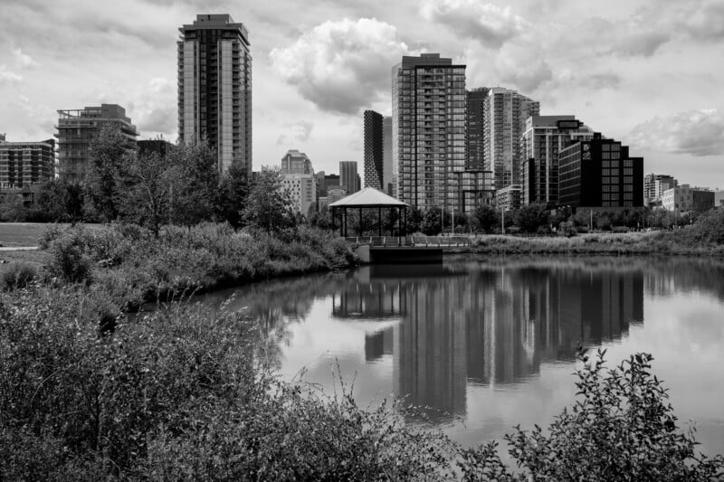 Black and white photo of a cityscape with tall buildings, a small pavilion on the water, and trees and shrubs surrounding a calm pond reflecting the structures and sky above.
