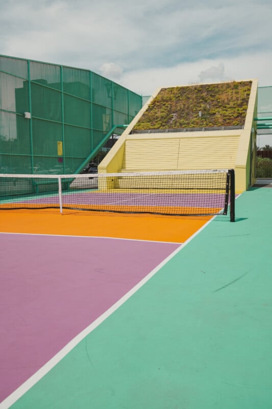 A colorful tennis court with purple, orange, and teal sections is set beside a green mesh fence and a unique structure with a slanted roof covered in grass and plants under a partly cloudy sky.