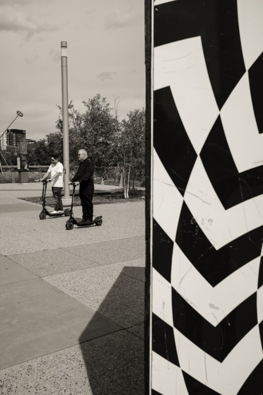 Two people stand on electric scooters in an outdoor area with trees and city buildings in the background. The right side of the image features a large black and white geometric mural in the foreground.