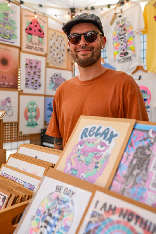 A smiling person in sunglasses and a cap stands behind a display of colorful, illustrated art prints and posters at a market stall, with vibrant artwork and T-shirts hanging in the background.