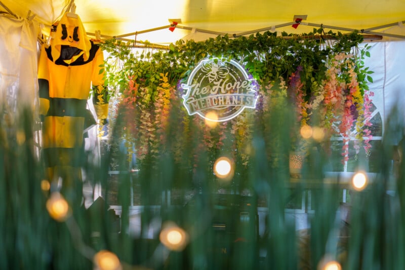 A yellow beekeeper suit hangs beside a neon sign reading "The Honey Merchants," with artificial greenery and colorful hanging flowers decorating the booth. Out-of-focus lights and plants appear in the foreground.