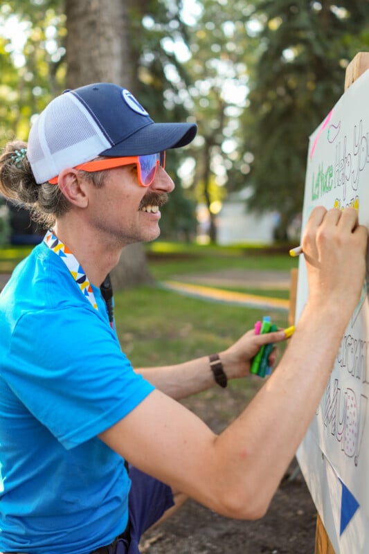 A person in a blue shirt, cap, and orange sunglasses writes on a whiteboard outdoors, holding colorful markers, with trees and sunlight in the background.