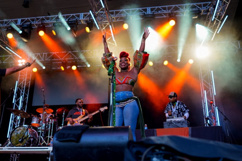 A singer with raised arms performs energetically on stage with a live band, colorful lights, and smoke effects creating a lively concert atmosphere.