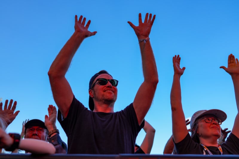 A group of people at an outdoor event raise their hands in the air. A man in sunglasses and a dark shirt is centered, smiling, and enjoying the moment, with a bright blue sky as the background.