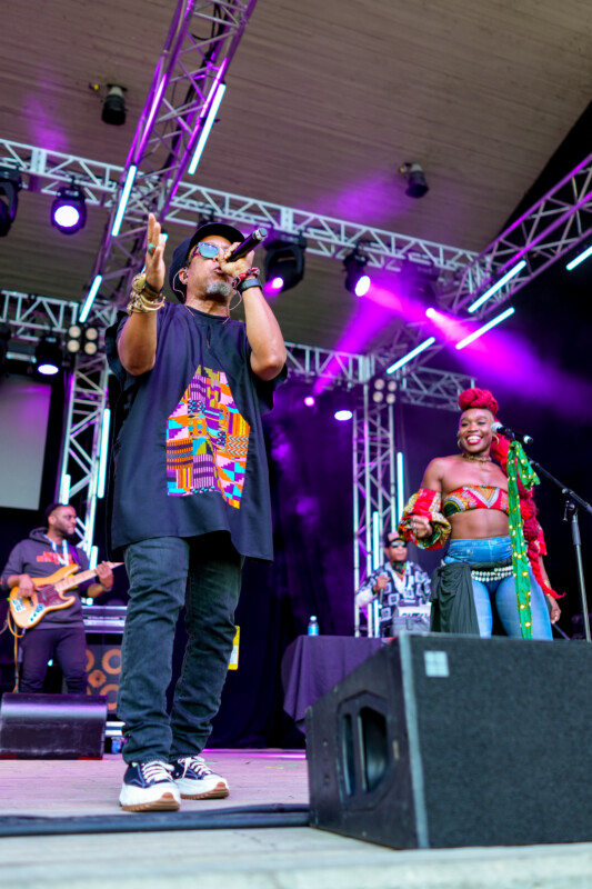 A male rapper in a colorful graphic shirt performs on an outdoor stage with a microphone, while a smiling female singer in vibrant attire stands beside him; band members play instruments in the background under purple stage lights.