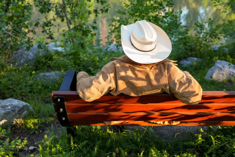 A person wearing a white cowboy hat and tan jacket sits on a wooden bench outdoors, surrounded by greenery and rocks, facing away from the camera toward a blurred natural background.