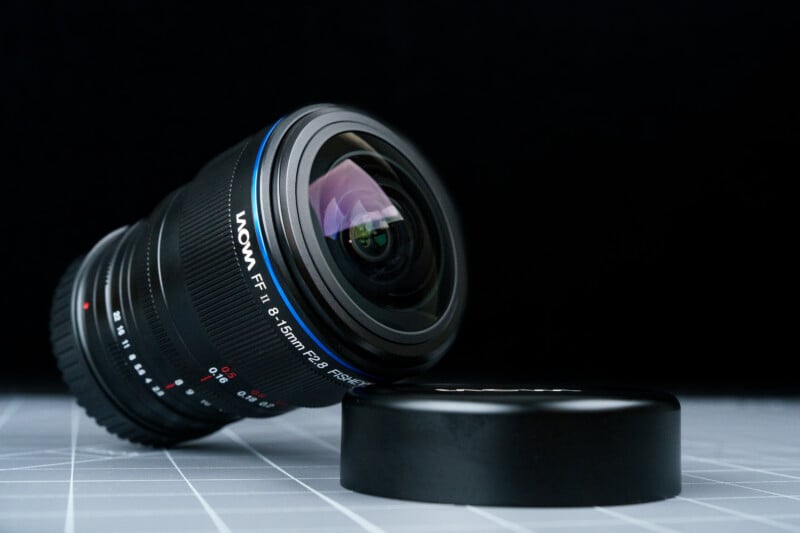 A Laowa camera lens with a blue accent ring is lying on its side on a grid-patterned surface, next to a detached lens cap. The background is black.