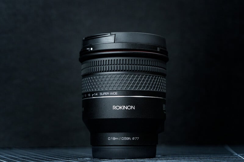 A black Rokinon camera lens stands upright on a dark, textured surface with a matching dark background. The lens has white text labels and visible grip ridges along the body.