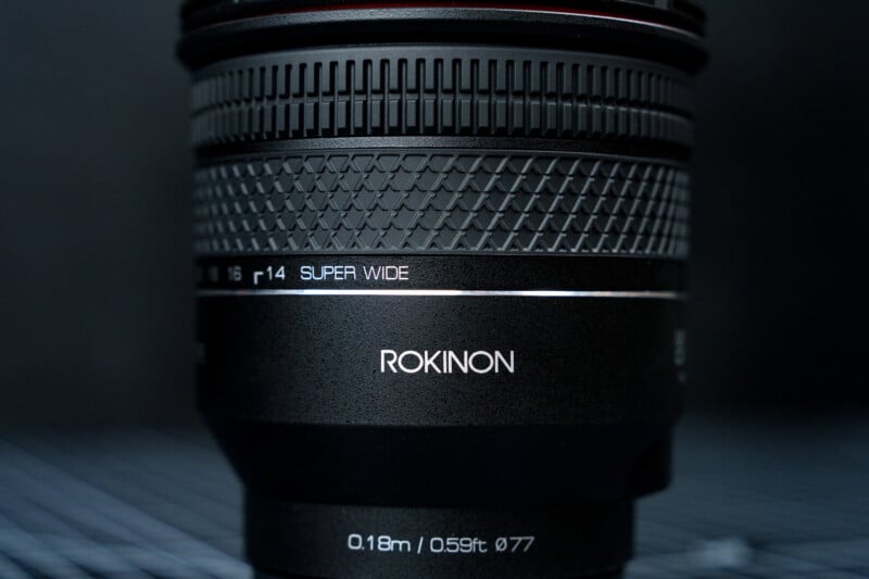 Close-up of a Rokinon camera lens showing detailed texture, engraved text reading "r14 SUPER WIDE" and "ROKINON," and specifications "0.18m / 0.59ft Ø77" on a dark, blurred background.
