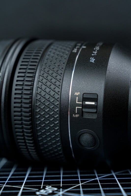 Close-up of a camera lens with a textured grip, showing a switch labeled AF/MF for selecting between autofocus and manual focus modes, against a dark background.