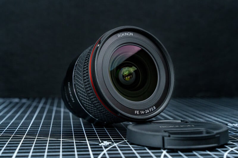 A Rokinon FE 14-24mm F2.8 camera lens is placed on a grid-patterned surface with its lens cap lying nearby. The background is dark and textured.