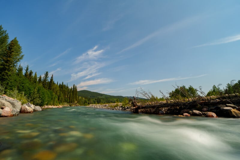 A clear river flows swiftly past rocks and fallen trees, surrounded by dense green forest and distant hills under a bright blue sky with wispy clouds.