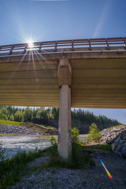 A sunburst appears above the railing of a concrete bridge over a river, with trees and shrubs along the riverbank under a clear blue sky.