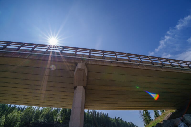 A view from beneath a concrete bridge on a sunny day, with the sun shining brightly above, creating lens flare. Trees and blue sky are visible in the background.