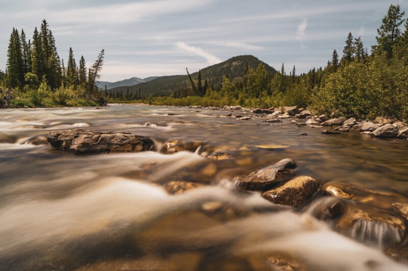 A scenic river flows over rocks in the foreground, surrounded by dense green trees and hills under a partly cloudy sky, with distant mountains in the background.