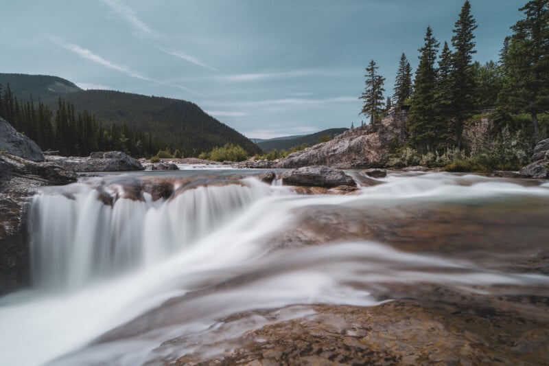 A scenic view of a small waterfall flowing over rocks, surrounded by evergreen trees and forested hills under a partly cloudy sky.