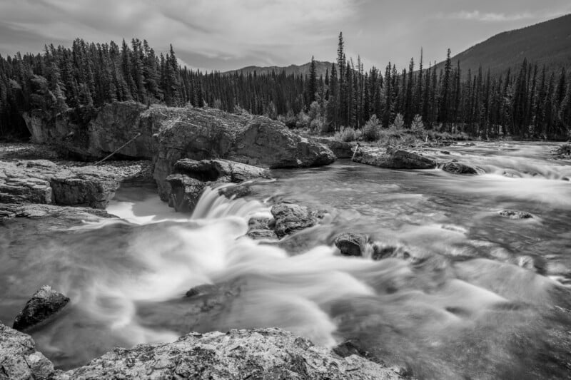 Black and white photo of a river with small waterfalls flowing over rocks, surrounded by dense pine forests and distant mountains under a partly cloudy sky.