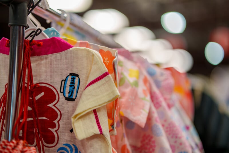 Colorful traditional Japanese children's yukatas with floral and lantern patterns hang on a rack, displayed in a well-lit setting with soft, blurred lights in the background.