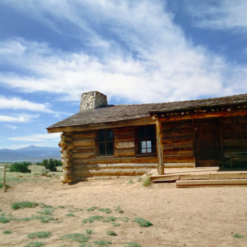 A rustic log cabin with a stone chimney stands on dry, sandy ground under a blue sky with wispy clouds. Mountains are visible in the background, and sparse vegetation surrounds the cabin.