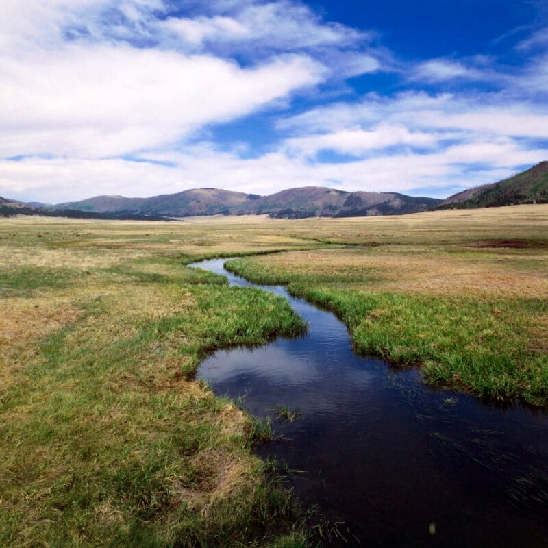 A small stream winds through a grassy meadow under a bright blue sky with scattered white clouds, with rolling hills and distant mountains in the background.