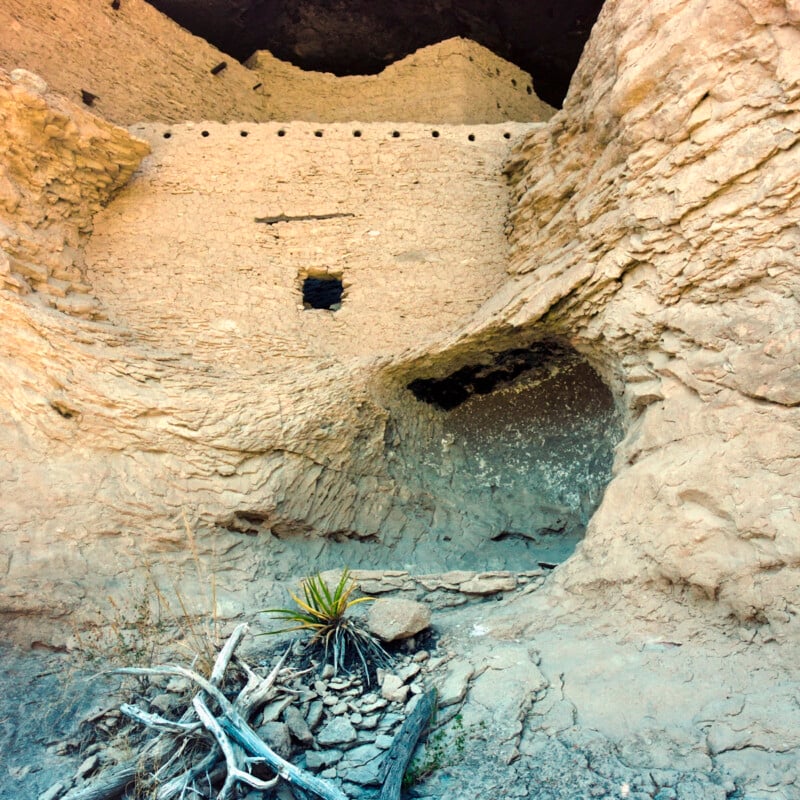 Ancient cliff dwelling built into a rocky sandstone cliff face, with small windows and holes visible in the structure. At the base, there are desert plants and dry fallen branches.