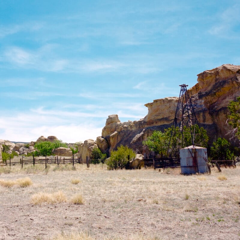 An old metal windmill structure stands next to a small shed amid dry grass and scattered bushes, with rocky cliffs and a wooden fence in the background under a blue sky.