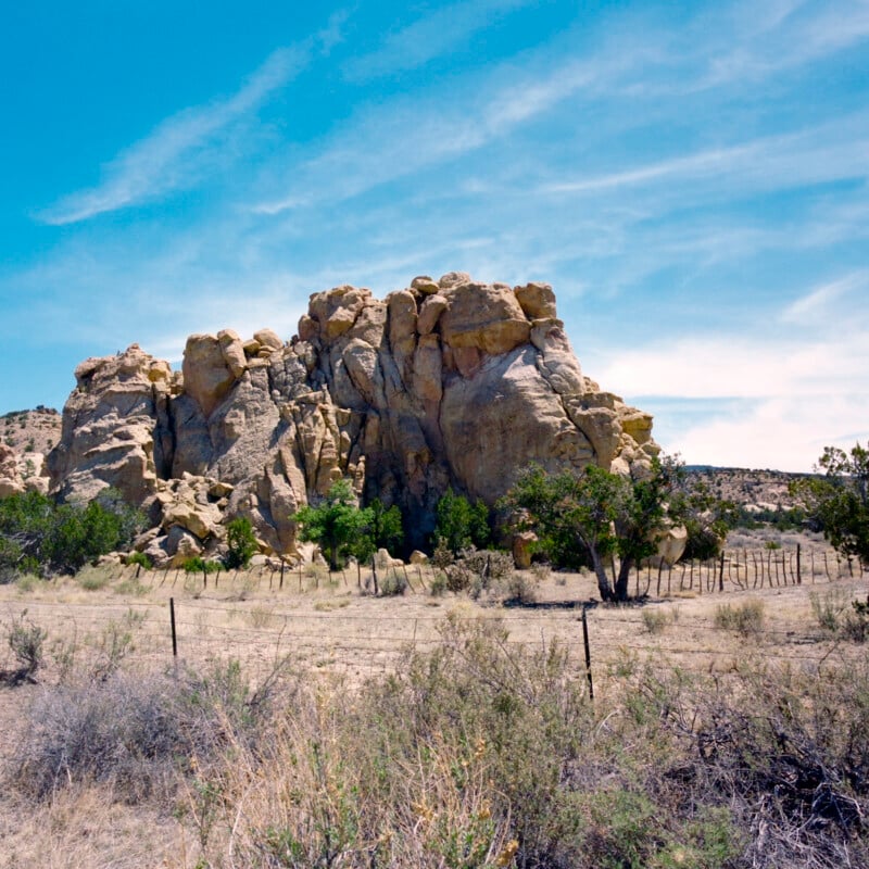 A large, rugged rock formation rises above dry grass and sparse shrubs under a bright blue sky with wispy clouds. Small trees grow near the base, and a wire fence runs along the foreground.
