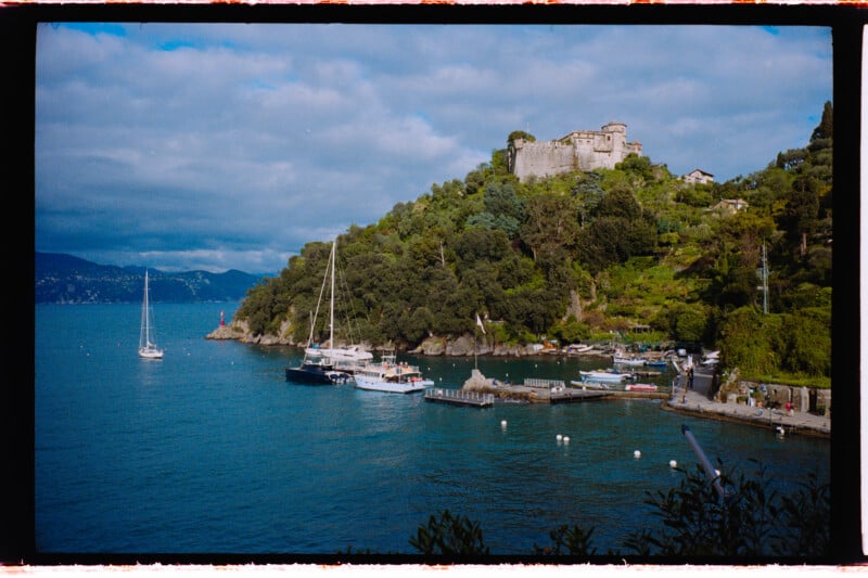 A scenic view of a small marina with sailboats docked beside a green hillside, topped by a historic stone fortress, under a cloudy blue sky by the sea.