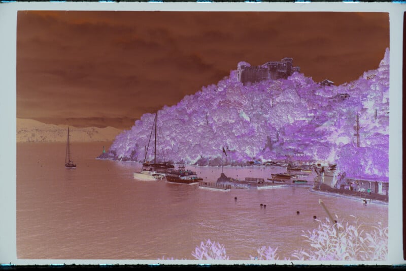 A color-inverted image of a seaside harbor with sailboats, a hilly shoreline, and a large building on the hilltop, under a cloudy sky. The colors appear purple, pink, and brown due to the negative effect.