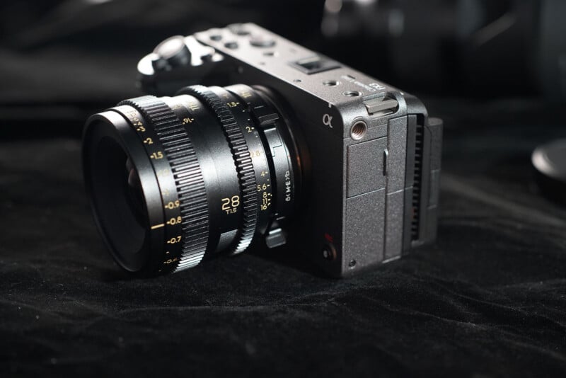 A close-up of a Sony Alpha digital camera with a black 28mm lens resting on a dark, textured surface. The lens features yellow focus and aperture markings.