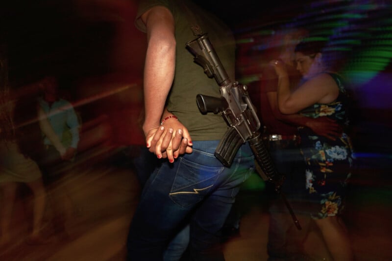 A person with a rifle slung over their shoulder holds hands with another person while dancing in a dimly lit, crowded room with colorful light streaks in the background.