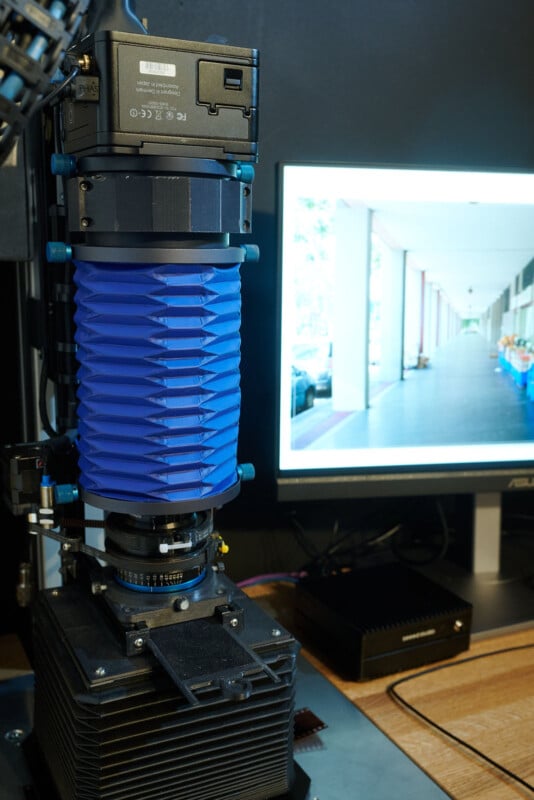 A close-up of a high-tech camera with a blue accordion-like part is set up on a desk, pointed at a monitor displaying a clear image of a building corridor with columns and parked cars.