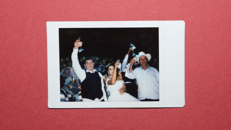 A printed photo shows three people dressed formally, raising glasses in a toast at a celebration, possibly a wedding, against a dark background. The photo is placed on a red surface.