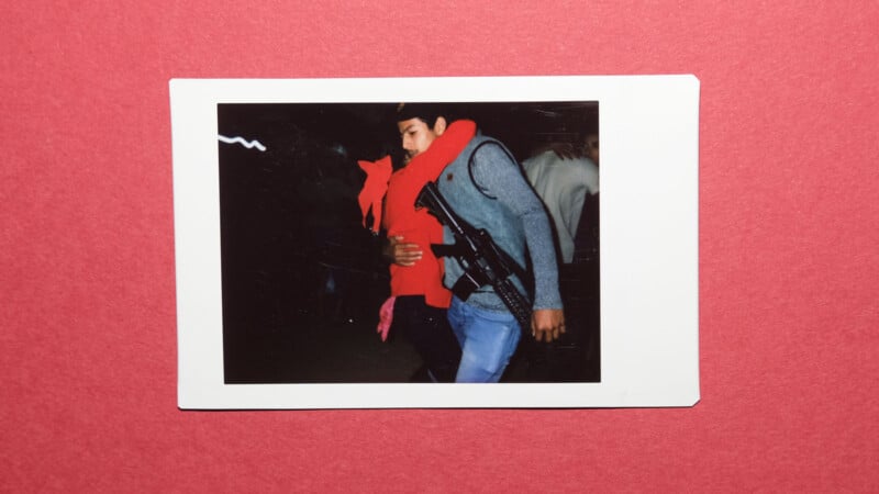 A photo of a person in a red hoodie being embraced or carried by someone in a gray jacket and blue jeans, set against a red background. The photo appears to be a printed instant film photograph.
