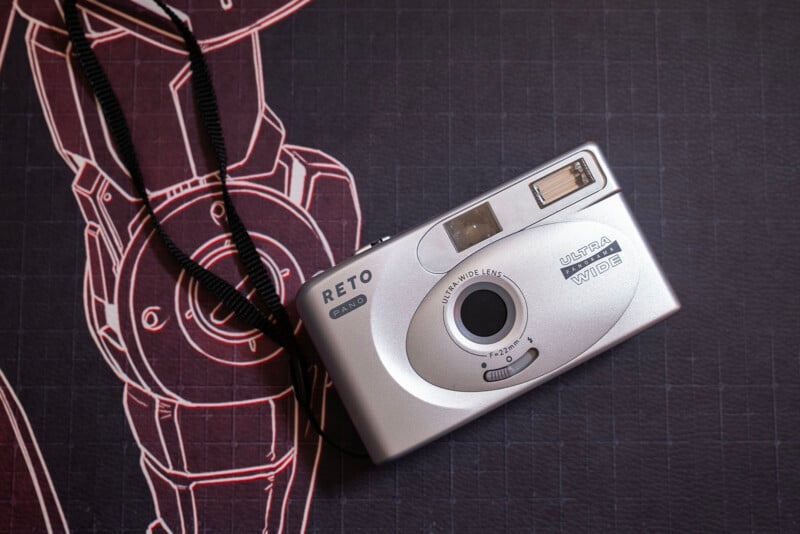 A silver RETO Ultra Wide & Slim film camera with a black wrist strap rests on a dark surface featuring a technical drawing of a robot in white and red lines.