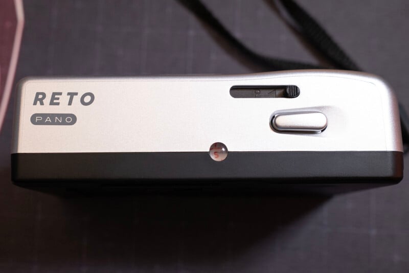 Top view of a silver and black RETO Pano film camera, showing the shutter button, a small slider switch, and part of a black camera strap on a dark surface.