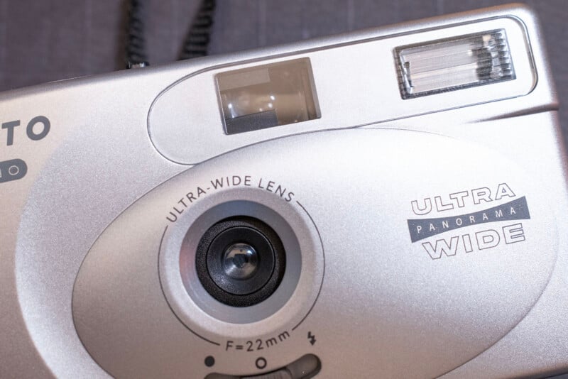 Close-up of a silver film camera featuring a prominent 22mm ultra-wide lens, viewfinder, and flash. The camera body displays the text "ULTRA WIDE" and "PANORAMA.