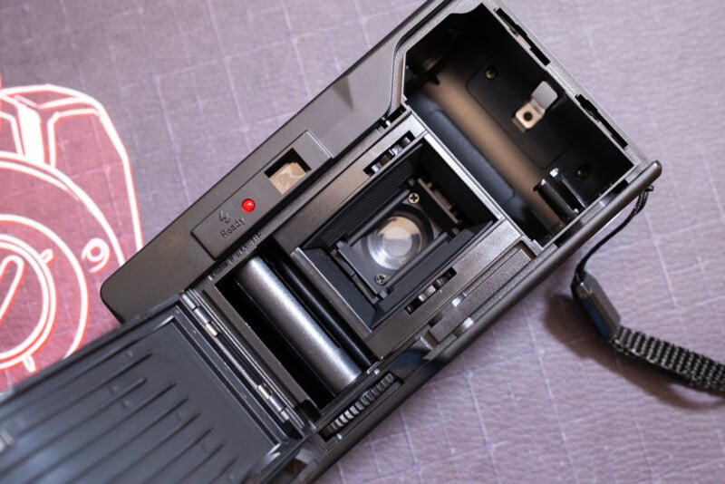 Open back of a film camera showing the film compartment, pressure plate, lens, and take-up spool, with a wrist strap attached and a red button visible beside the film chamber. The camera rests on a textured surface.