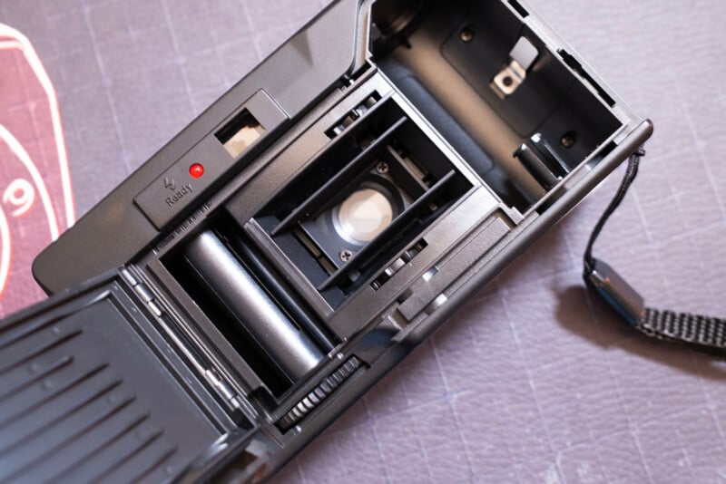 Close-up of an open film camera showing the film compartment, lens, film spool area, and a wrist strap on a textured surface.