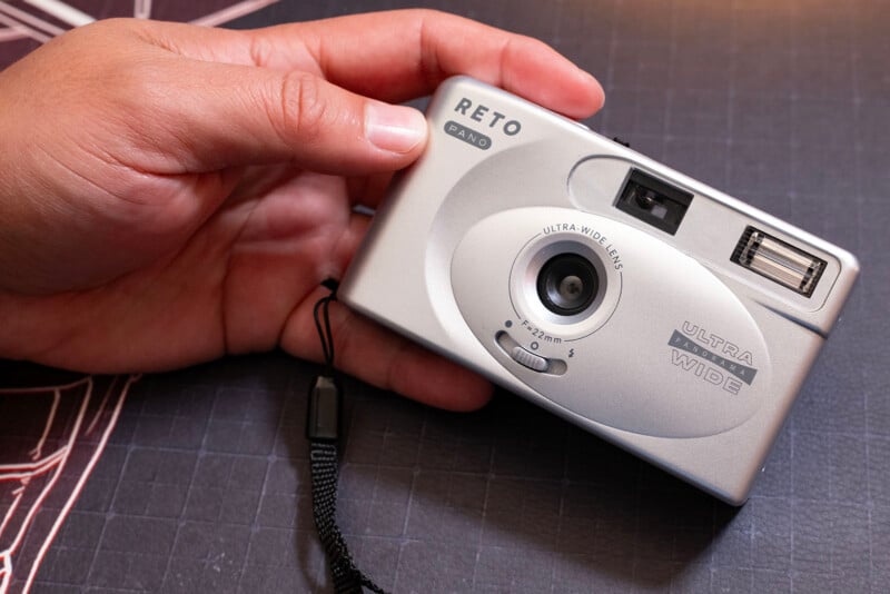 A hand holding a silver RETO Ultra Wide and Slim 35mm film camera with a wrist strap, over a dark textured surface.
