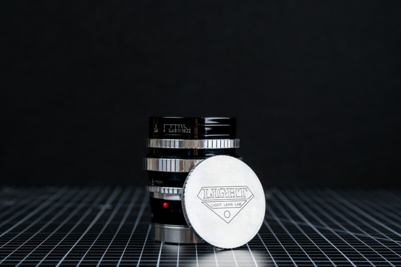 A camera lens with metallic rings stands on a grid-patterned surface. In front of it, a silver lens cap with the word 