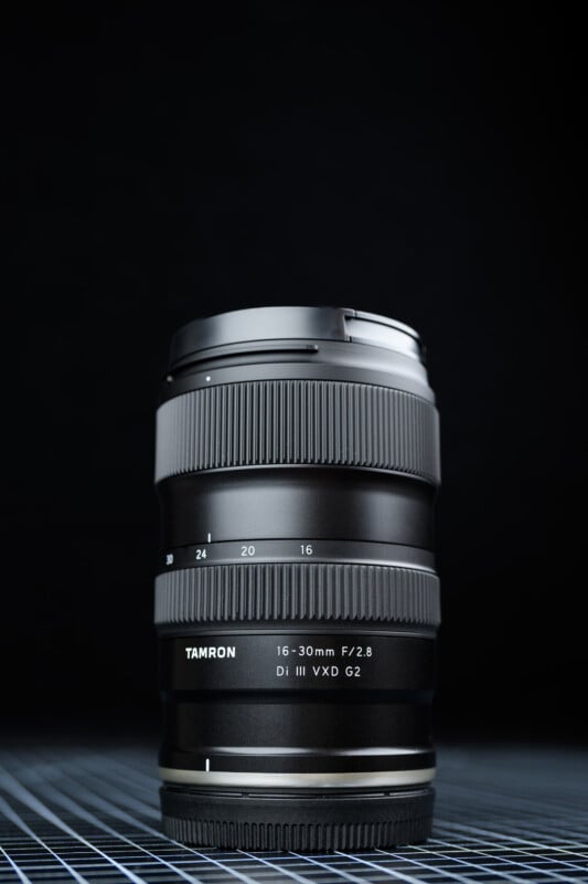 A Tamron 16-30mm F/2.8 Di III VXD G2 camera lens stands upright on a gridded surface, with a dark, blurred background.