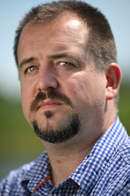 A man with light skin, short brown hair, and a goatee gazes at the camera. He is wearing a blue and white checkered shirt, standing outdoors with a blurred green background.