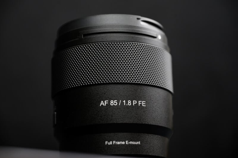 A close-up photo of a black camera lens labeled "AF 85 / 1.8 P FE" with a textured grip and "Full Frame E-mount" written near the base, set against a dark background.