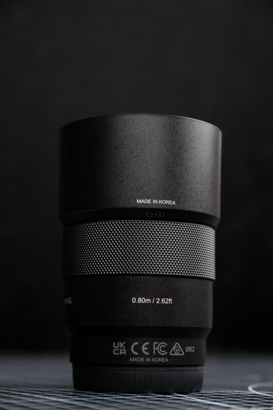 A close-up of a camera lens standing upright on a surface, with labels such as "MADE IN KOREA" and focus distance markings visible against a dark, blurred background.
