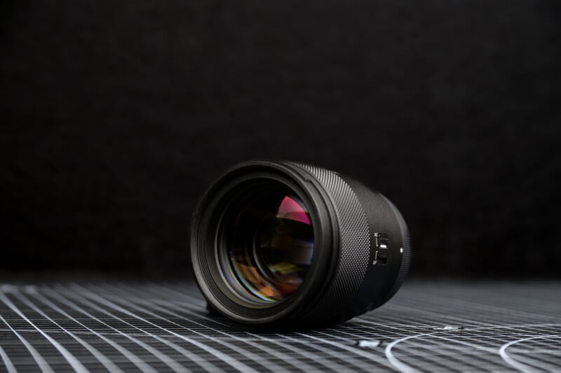 A camera lens with a textured focus ring lies on its side on a grid-patterned surface, with a dark, blurred background.