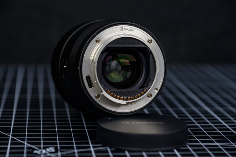 A camera lens lies on its side on a grid-patterned surface, with the rear glass element and electronic contacts visible. The lens cap labeled "SAMYANG" rests nearby, slightly out of focus.
